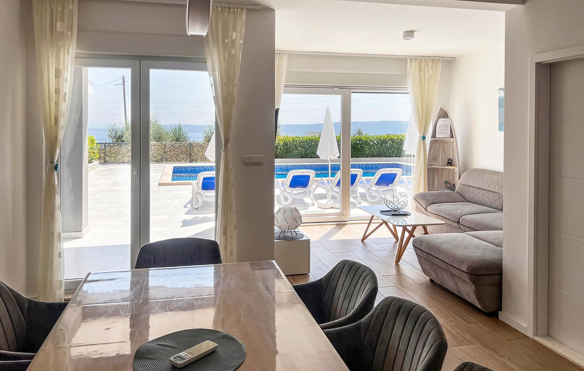A modern living room with a glass door leading to a pool area. There is a dining table with chairs, a sofa, and a small side table.