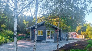 A small cabin with a gravel driveway, surrounded by trees and a wooden deck.