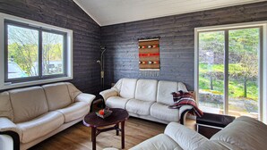 A living room with a dark wooden wall, a white sofa, a smaller white sofa, a coffee table, and a large window with a view of trees and a car outside.