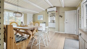 L’intérieur chaleureux d’un chalet avec une table à manger en bois, des chaises blanches, un coin cuisine doté de meubles blancs et une grande fenêtre laissant entrer la lumière naturelle.