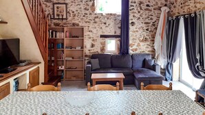 A cozy living room with a stone wall, a dark sofa, a wooden bookshelf, a television, and a dining area with a patterned tablecloth.