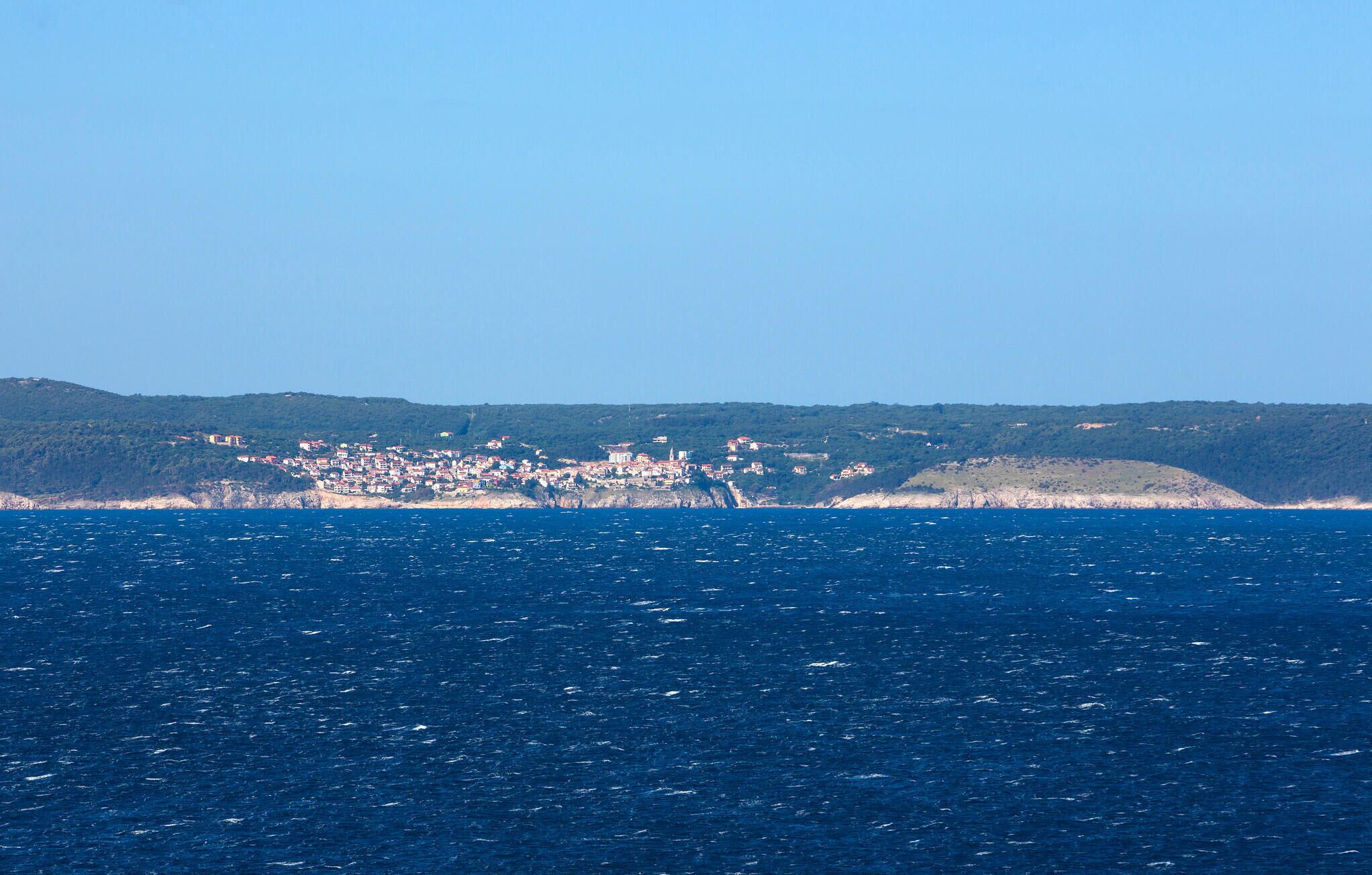 En kystby med bygninger langs kystlinjen, en klar blå himmel og udsigt over havet.
