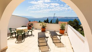 A balcony with a table and chairs, potted plants, and a view of the sea and mountains.