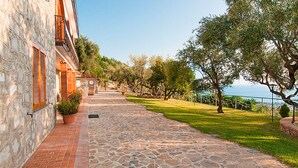 A stone pathway leading to a building with a balcony, surrounded by greenery and a view of the sea.