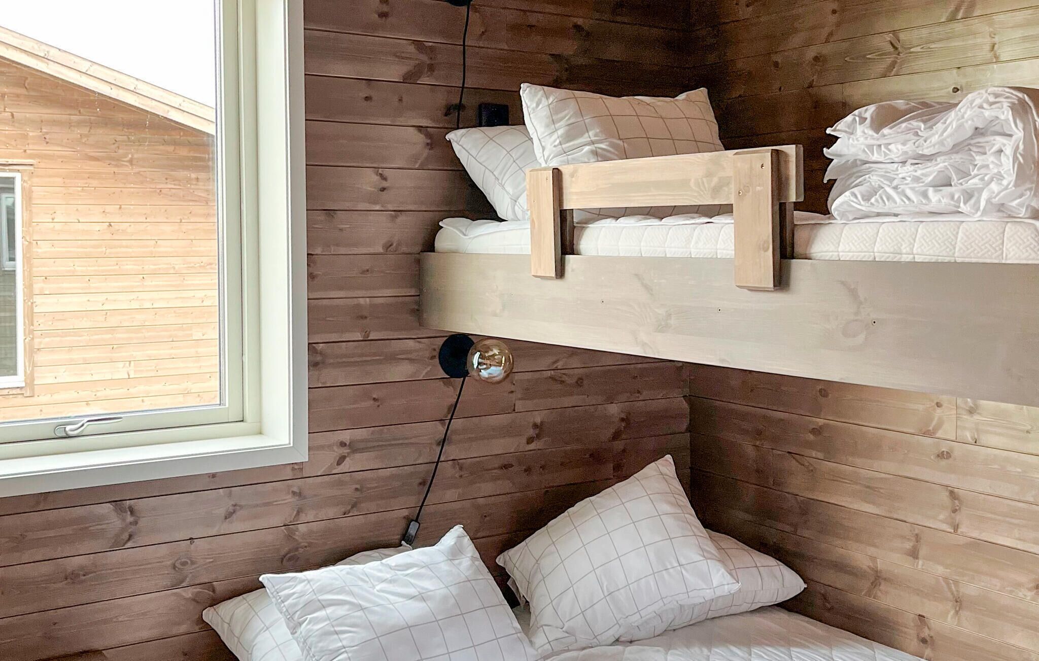 En køjeseng med træramme og hvidt sengetøj, et vindue med udsigt til en træbygning, og en hængende lampe.