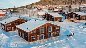 En grupp sammanbyggda trähus med stora fönster, omgivna av snötäckt mark och träd, vilket tyder på att miljön är vinterlig.