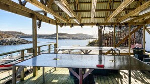 Um deck de madeira com mesa de metal, com vista para um litoral rochoso e mar calmo.