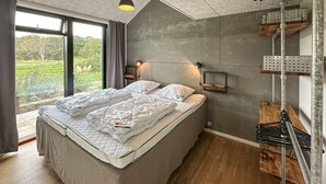 A bedroom with a large bed, a view of greenery outside, and a concrete wall with a metal shelf. - 3 bedroom amazing home in Nexø (Nexø)