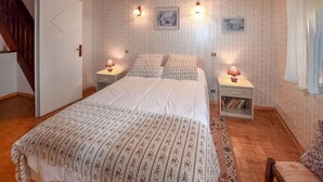 Un dormitorio con una cama, dos mesitas de noche, una mesita auxiliar, una puerta y una ventana con cortinas.
