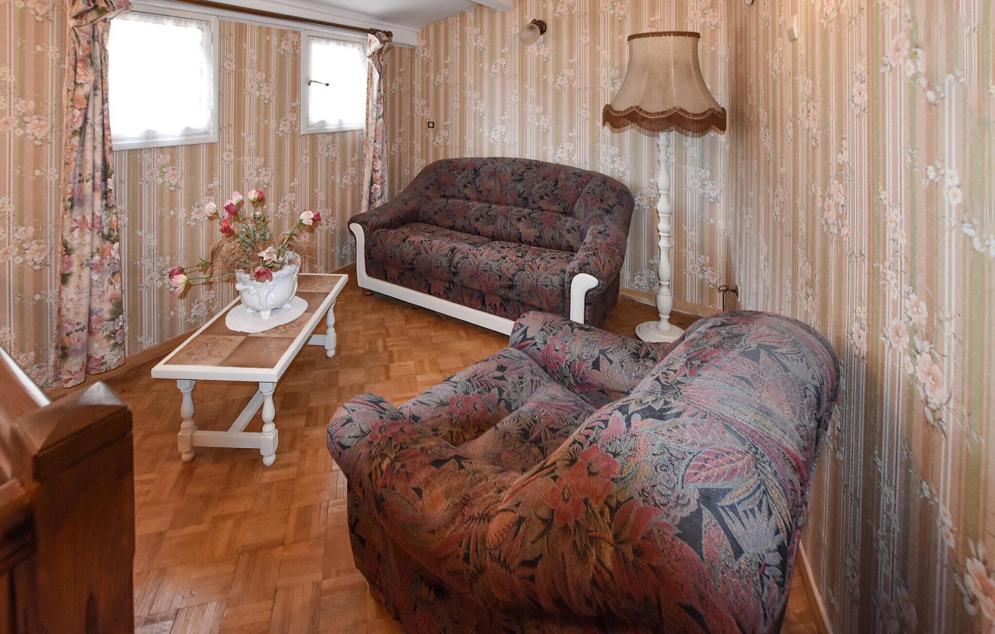 Une pièce comprenant un canapé à motifs floraux, un fauteuil blanc, une table basse en bois ornée d’un vase de fleurs et une lampe.