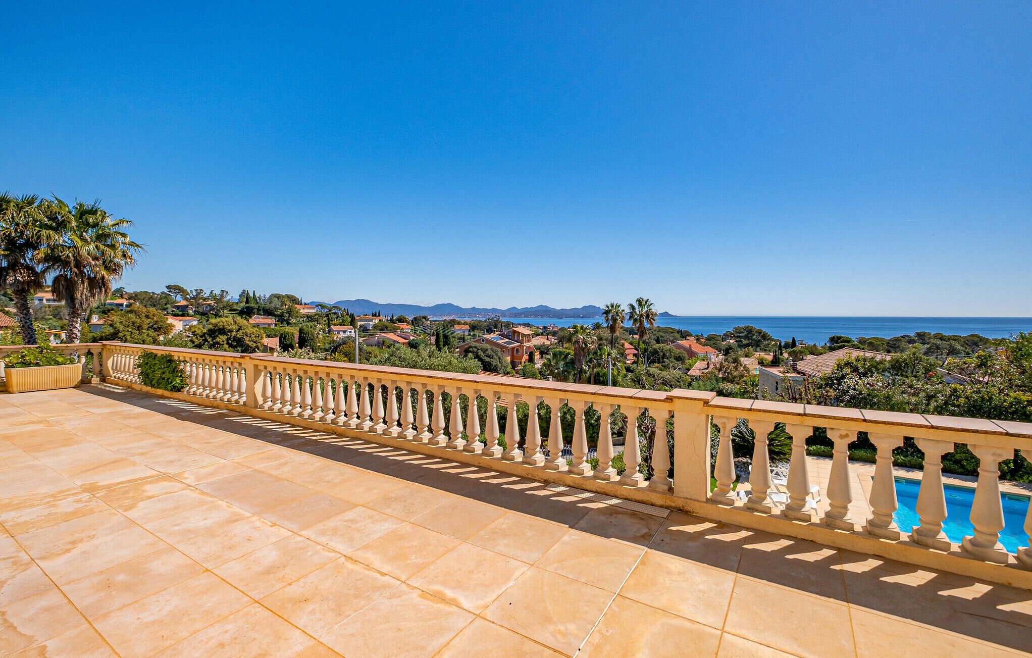 A terrace with a tiled floor and a balustrade overlooking a coastal town with houses and a clear blue sky.