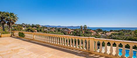 A terrace with a tiled floor and a balustrade overlooking a coastal town with houses and a clear blue sky.
