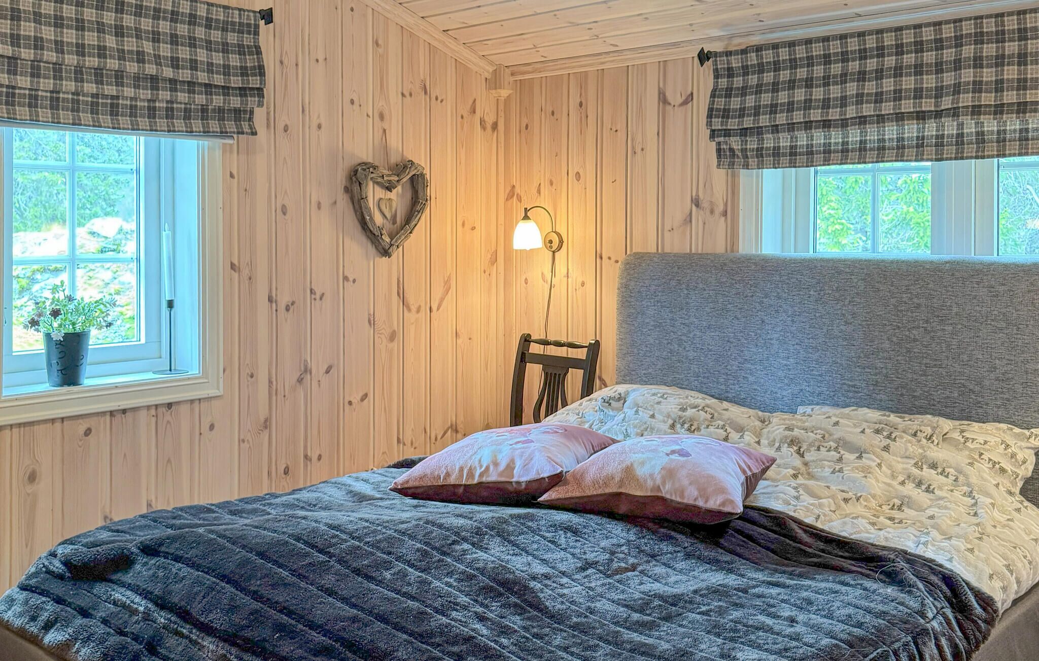 A bedroom with a wooden paneled wall, a heart-shaped decoration, a bedside lamp, a window with a view, and a bed with a quilt and pillows.