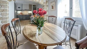 A dining area with a round wooden table, metal chairs, and a vase with flowers. There is a kitchen in the background with a stone wall and a window with curtains. - Gorgeous home in Roquemaure (Roquemaure)