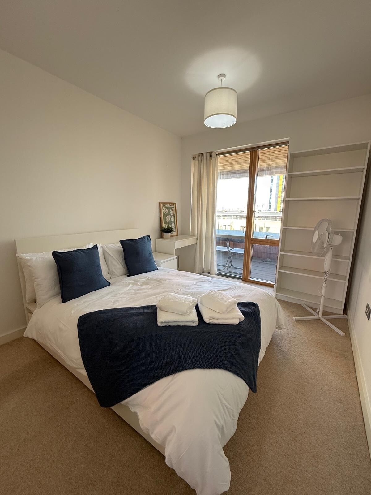 A bedroom with a bed, a bedside table, a nightstand, a window with curtains, and a standing fan.