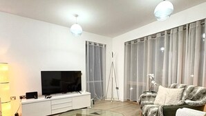A modern living room with a glass coffee table, a flat-screen TV, a sofa, and a large window with curtains. -  2-bedroom apartment near Canning Town Station E16 1GW London (London)