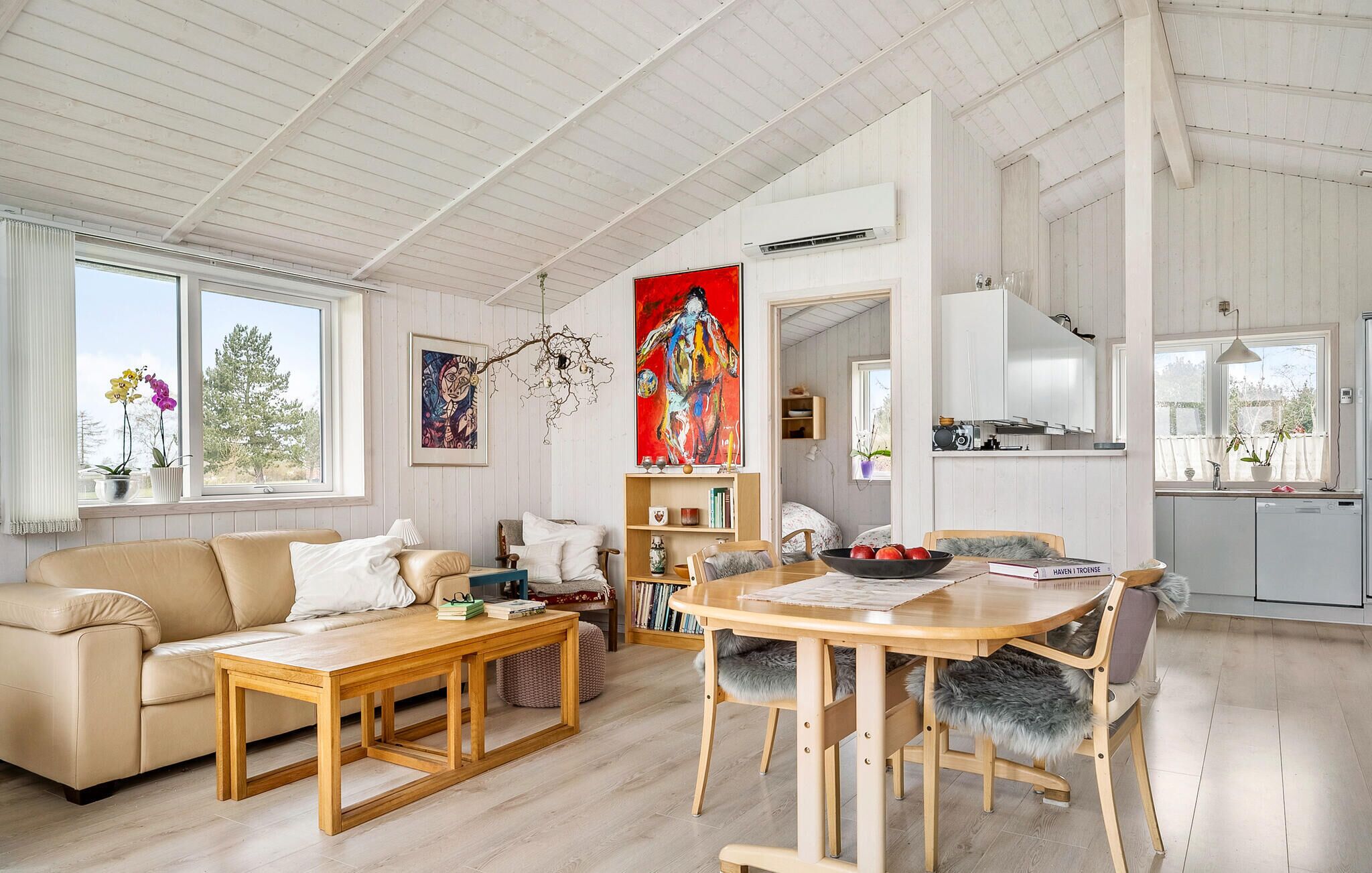 En rummelig stue med sofa, sofabord og spisebord. Der er et bogreol, et maleri og et køkkenområde at se.