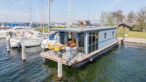 A floating houseboat docked at a marina with a small deck, a seating area with a table and chairs, and a lifebuoy on the deck.