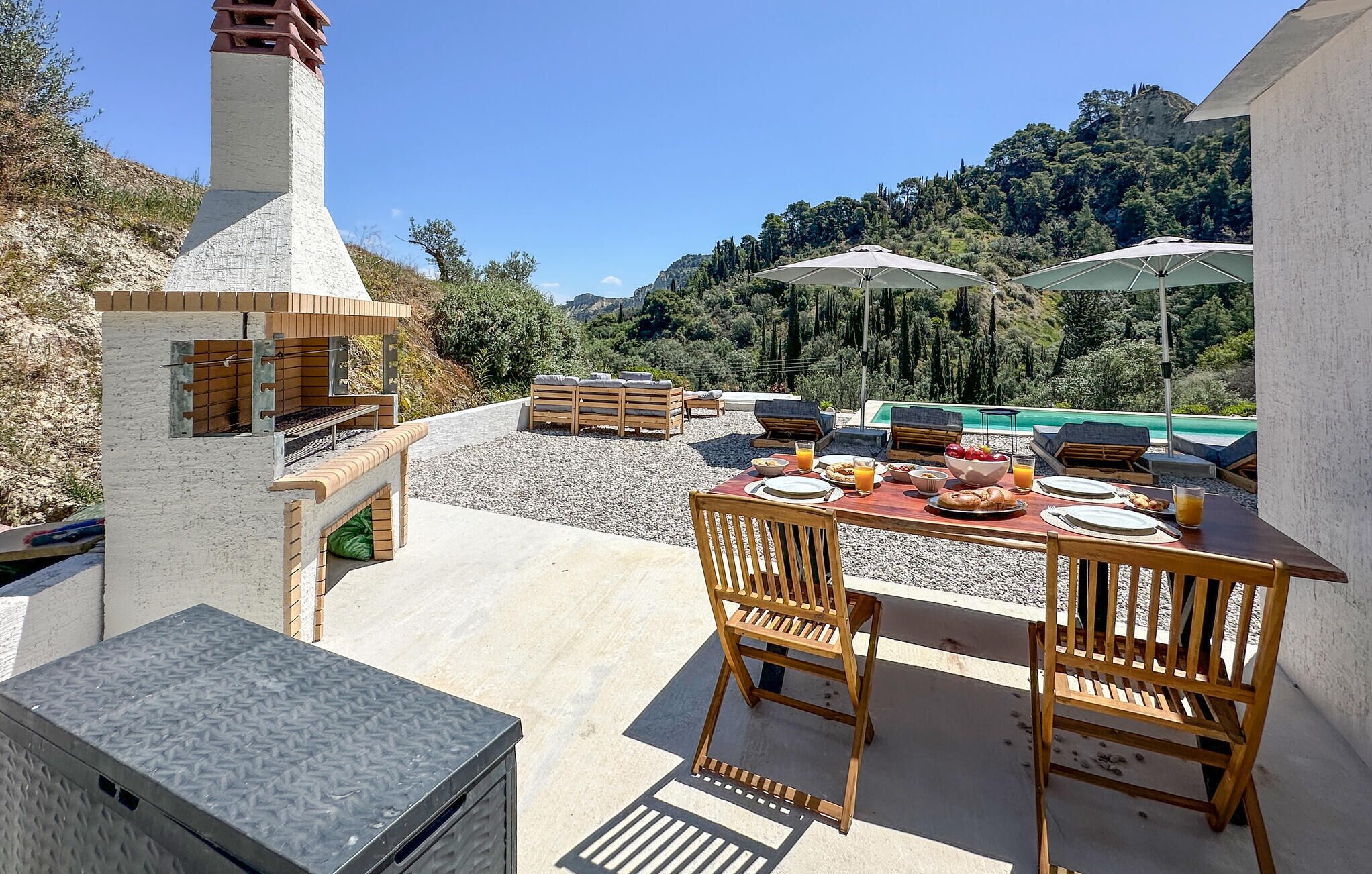 An outdoor dining area with a table set for breakfast, surrounded by wooden furniture and umbrellas, overlooking a pool and a mountainous landscape.