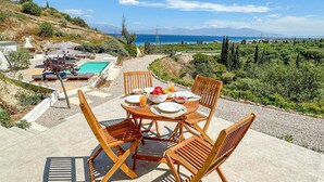 A dining table with chairs, a bowl of fruit, and a glass of juice on a patio overlooking a pool and a scenic view of the sea and hills.