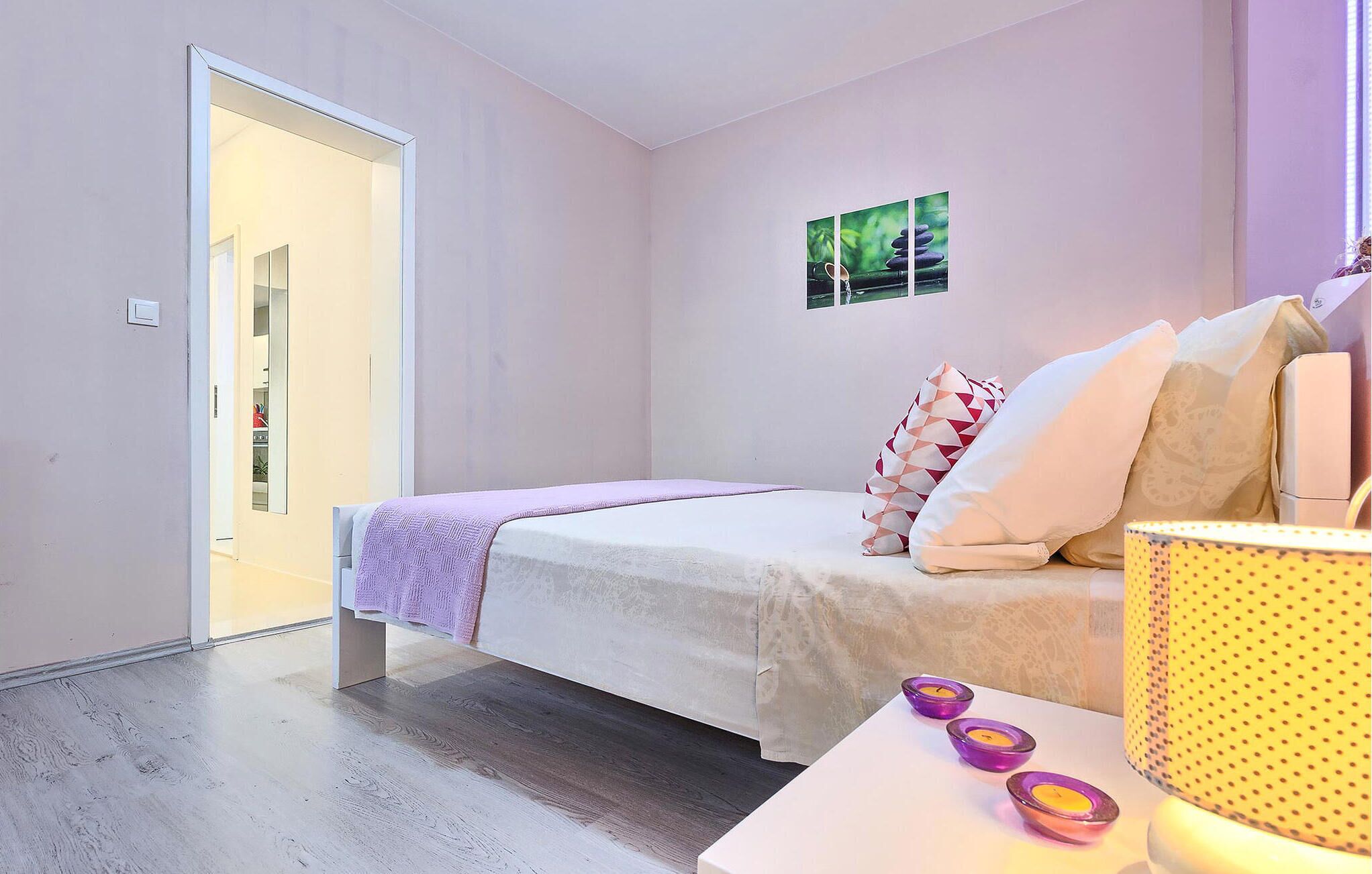 Ein Schlafzimmer mit Bett, Kissen, einem Nachttisch mit Kerzen und einem Fenster mit einem Gemälde.