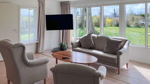 A living room with a sofa, two armchairs, a coffee table, a TV, and a view of a garden through large windows. - Lovely home in Kirke Hyllinge (Kirke Hyllinge)