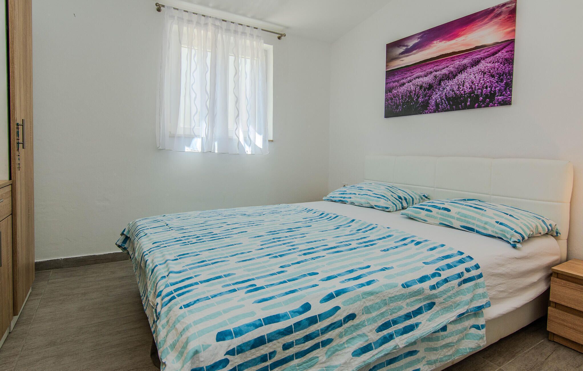 A bedroom with a bed, a window with sheer curtains, a nightstand, and a wall-mounted painting.
