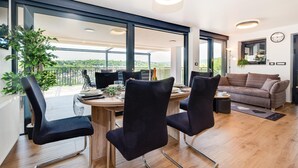 Ein modernes Wohnzimmer mit einem Holzesstisch, schwarzen Polsterstühlen, einem grauen Sofa und einem Balkon mit Blick auf Grünflächen.