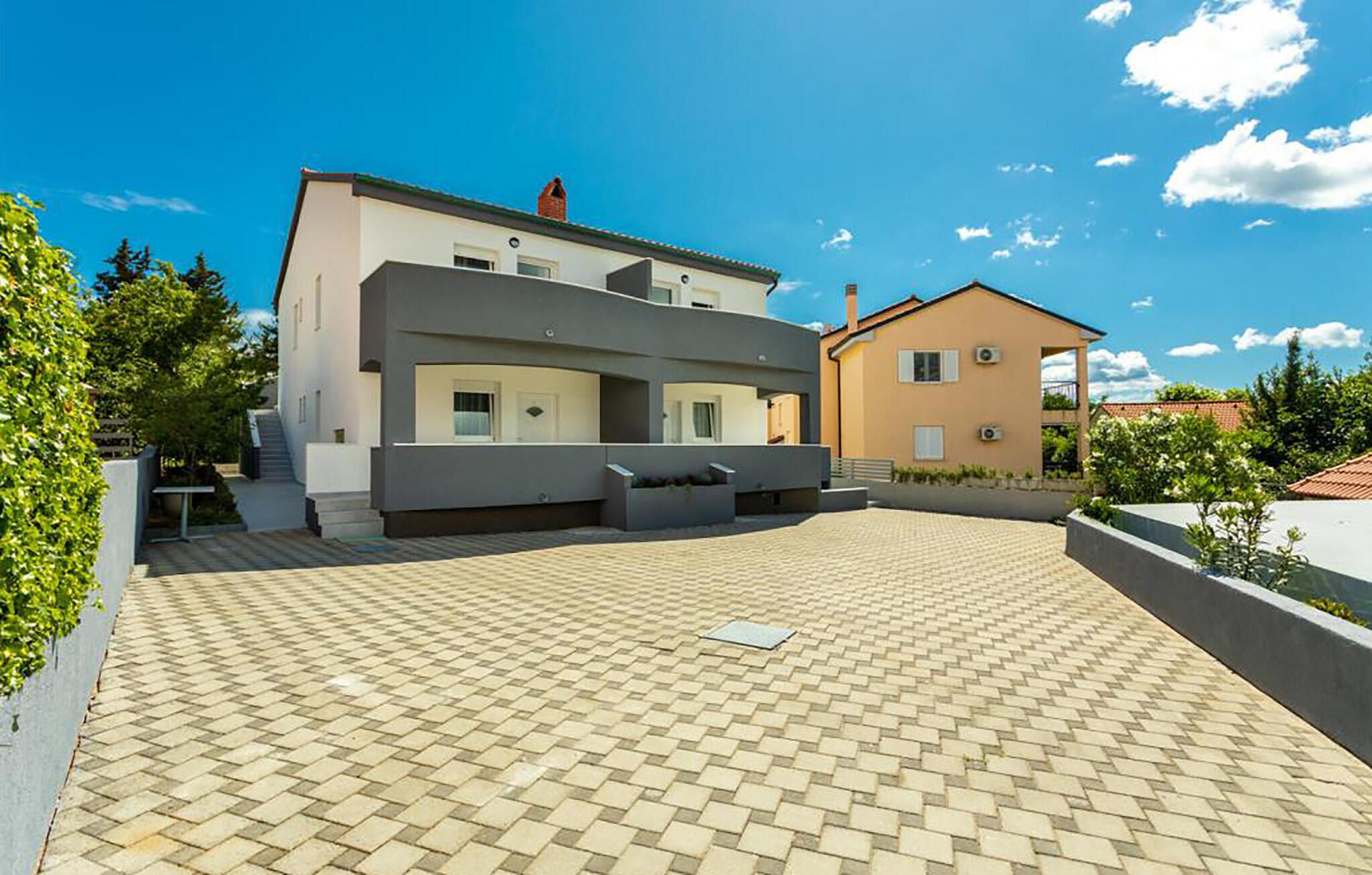 Une maison moderne à deux étages, dotée d’une cour dallée, d’un balcon et d’un petit jardin.