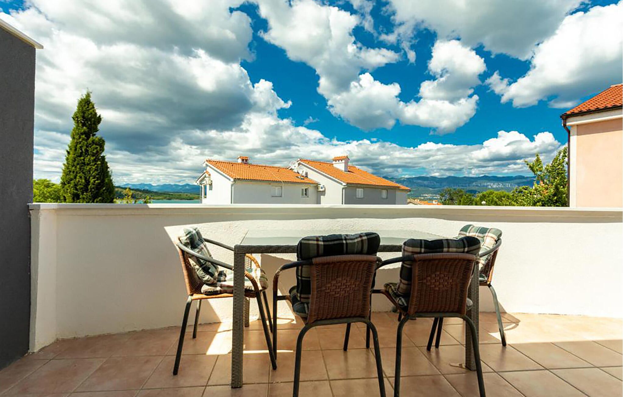 Une terrasse avec une table et quatre chaises en osier, donnant sur un quartier résidentiel avec des maisons et des montagnes, sous un ciel partiellement nuageux.