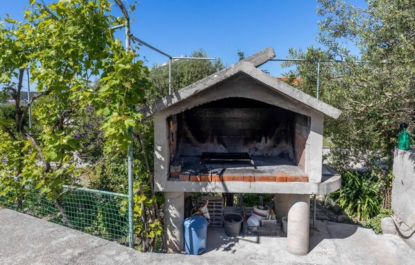 A concrete outdoor structure with a fireplace, surrounded by greenery and a metal fence.