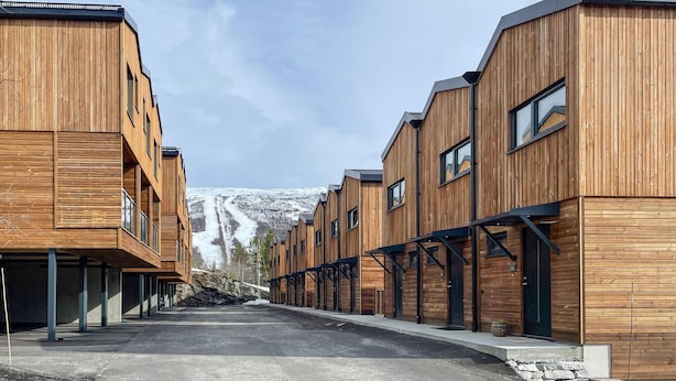 A row of modern wooden buildings with large windows and a view of a snowy mountain in the background.