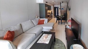 A modern living room with a grey sofa, a coffee table, and a TV stand. There is a dining area with a table and chairs in the background.