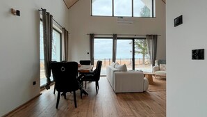 Una habitación amplia con suelo de madera, una zona de comedor con mesa y sillas, un sofá y ventanas grandes que ofrecen vistas al exterior.