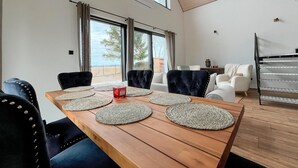 Una sala de estar moderna con una mesa de comedor de madera, sillas tapizadas oscuras y un sofá blanco. Amplias ventanas ofrecen vistas al exterior.