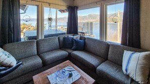 Una sala de estar acogedora con un sofá seccional gris, una mesa de centro de madera y grandes ventanales con vistas a un lago y montañas.