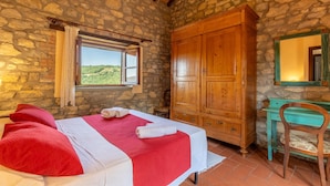 A bedroom with a stone wall, wooden wardrobe, a bed with red and white bedding, a window with a view, and a turquoise chair. - Nice home in Monte S.Maria Tiberina (Monte S.Maria Tiberina)