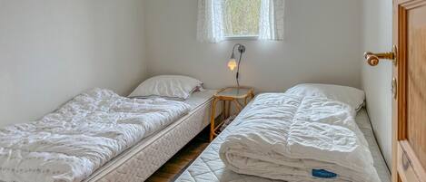 Et lille, rent værelse med to enkeltsenge, et natbord med lampe, og et vindue med gardiner.
