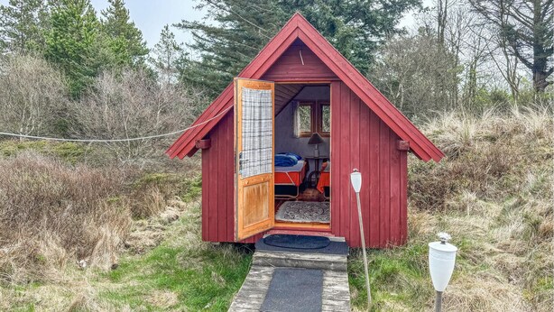 A small, red wooden cabin with a single bed and a window, surrounded by natural vegetation.