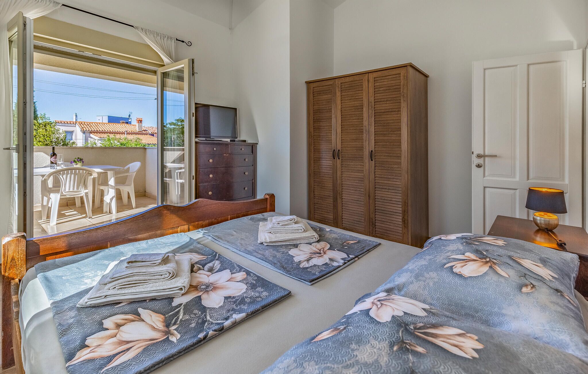 Une chambre à coucher comprenant un lit, une armoire en bois, un téléviseur posé sur une commode et un balcon aménagé avec du mobilier d’extérieur.