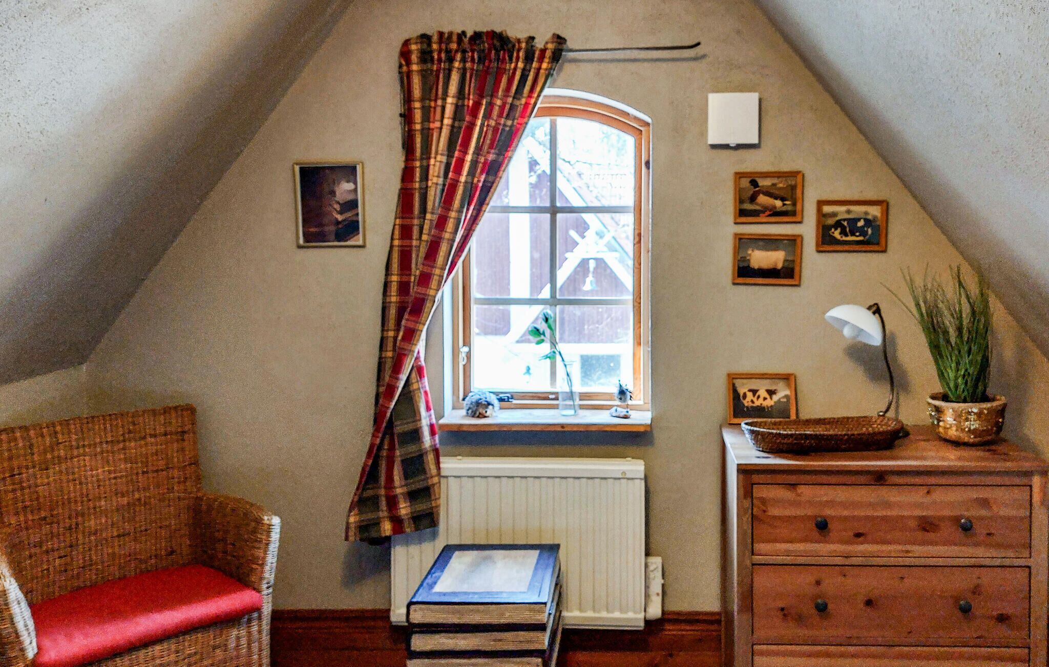 A cozy attic room with a sloped ceiling, a window with plaid curtains, a radiator, a wicker chair with a red cushion, a wooden dresser, and several framed pictures on the walls.