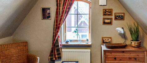 A cozy attic room with a sloped ceiling, a window with plaid curtains, a radiator, a wicker chair with a red cushion, a wooden dresser, and several framed pictures on the walls.