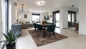 A modern living room with a sofa, a dining table with chairs, a TV mounted on the wall, and a large window. - Lovely home in Galizana (Galizana)