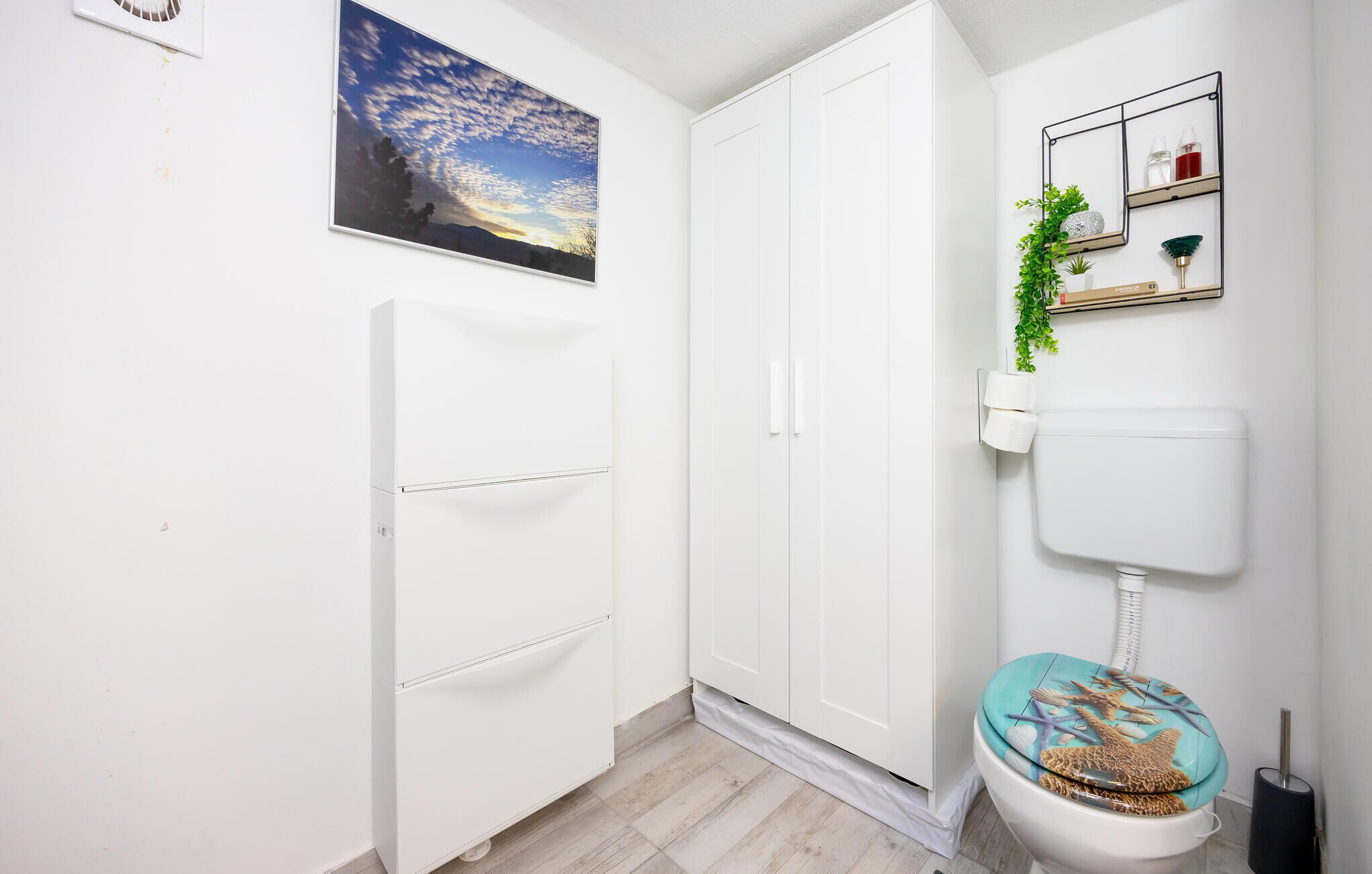 Ein Badezimmer mit Toilette, einer weißen Tür, einem wandmontierten Spiegel und einem Bilderrahmen mit Sonnenuntergangsmotiv.