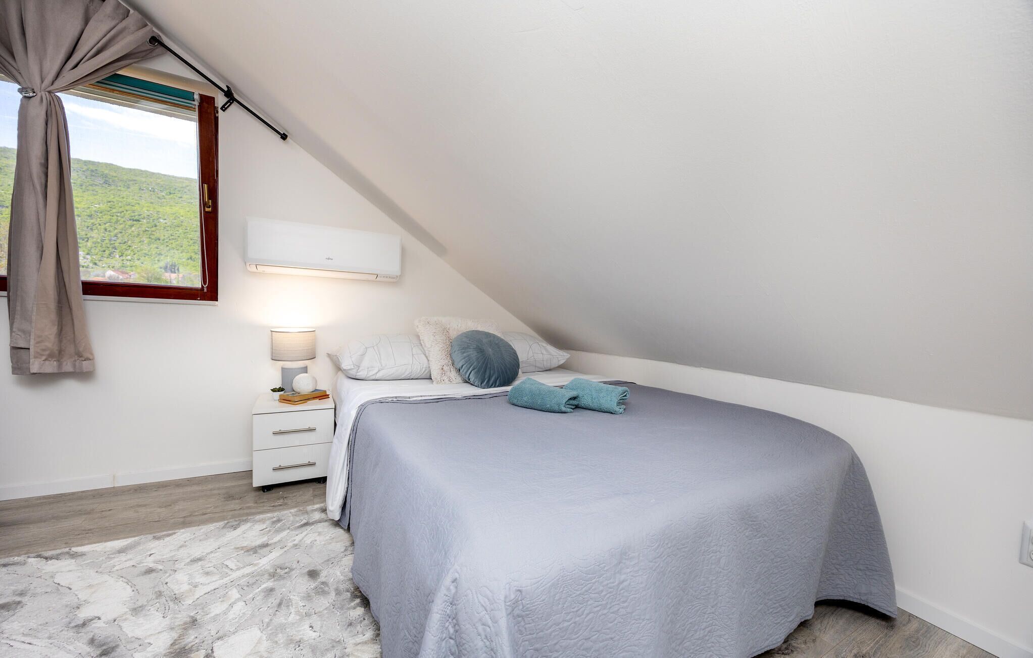 Une chambre avec un plafond en pente, un lit recouvert d’une couverture grise, une table de chevet avec une lampe, et une vue sur la verdure à l’extérieur.