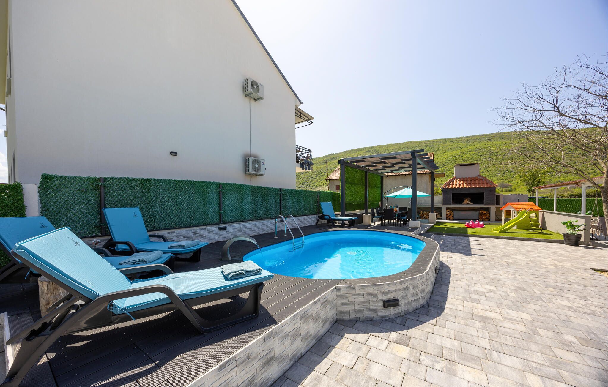 Piscina con sillas de descanso, zona de barbacoa y vista a una colina.