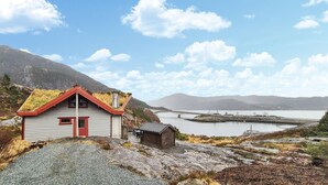 A small cabin with a sloped roof and a red door, situated on rocky terrain with a view of a lake and mountains in the background. - Beautiful home in Masfjordnes with sauna (Masfjordnes)