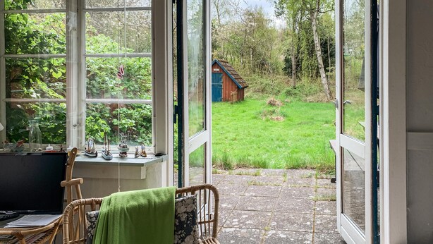 A room with a large window, a wicker chair with a green towel, a small table with a laptop, and a view of a garden with a wooden structure. - Cozy home in Knebel with WiFi (Knebel)