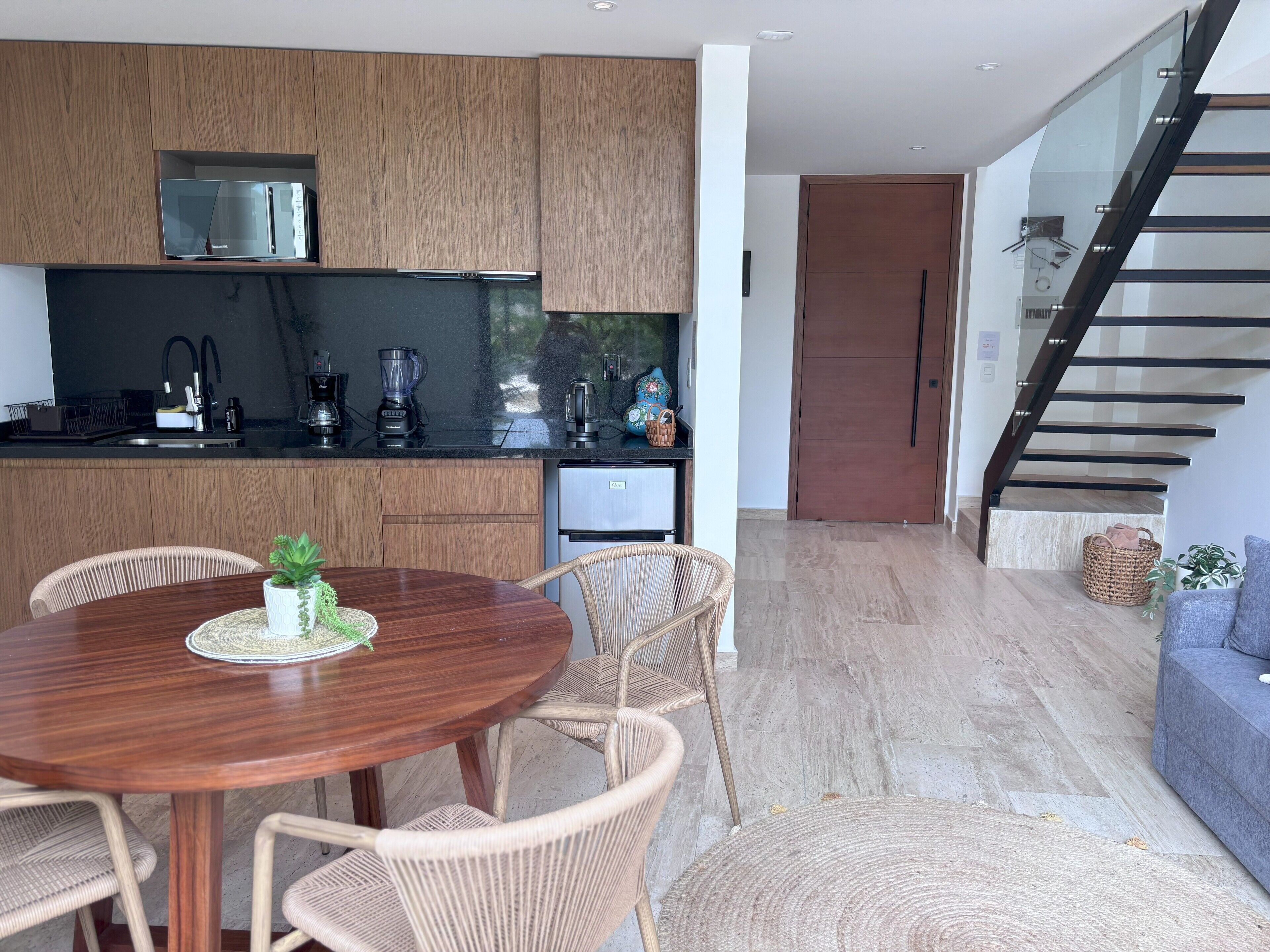 A modern kitchen with wooden cabinets, a built-in microwave, and a coffee maker. There is a dining area with a round wooden table and wicker chairs. A staircase with glass railings leads to another level.