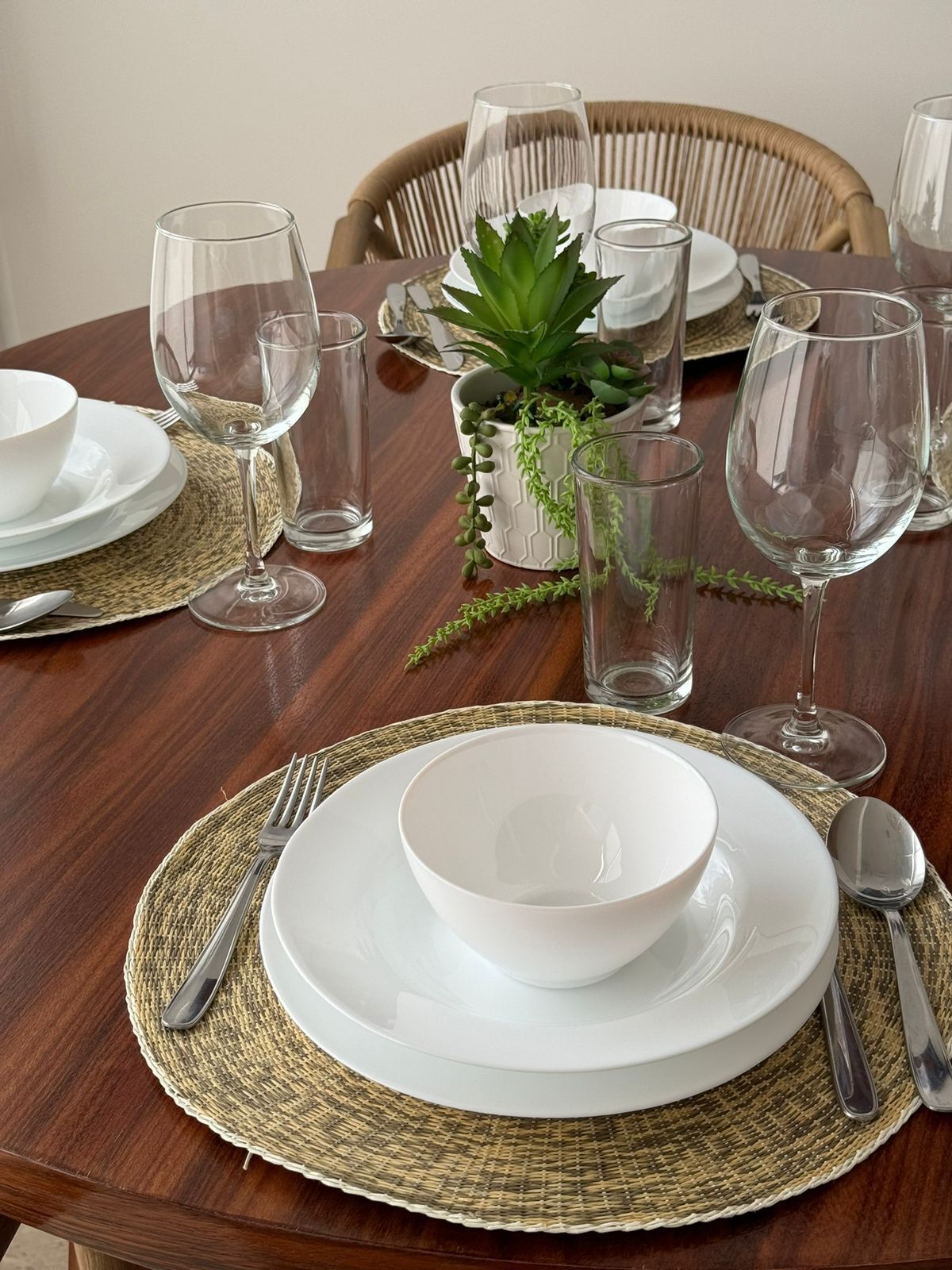 A dining table set with white plates, bowls, and cutlery, accompanied by clear glasses and a potted plant.